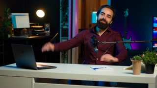 Happy influencer using microphone, arriving in dimly lit home studio interior, chatting with fans. Smiling man surrounded by podcast